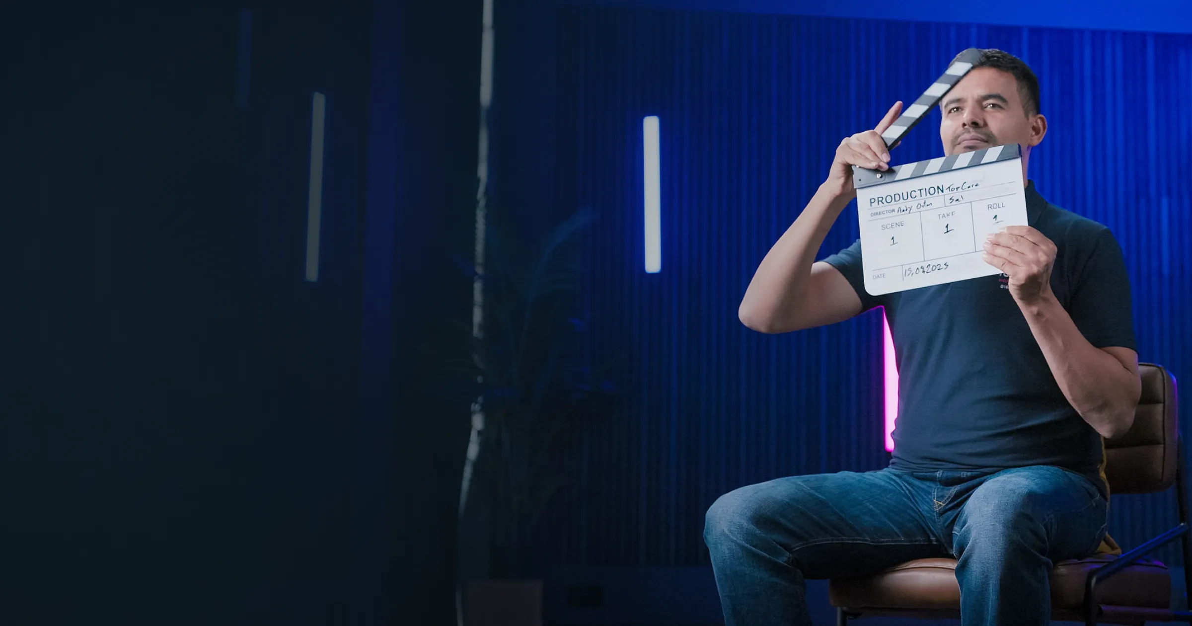 Man in a dark shirt sits on a chair, holding a clapperboard against a neon-lit blue background, suggesting a calm, professional atmosphere.